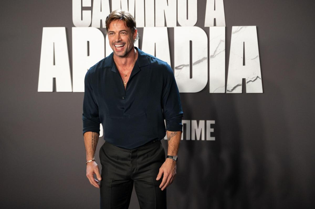 actor care poartă pantaloni și cămașă neagră și zâmbește în timp ce stă cu spatele la un panou publicitar