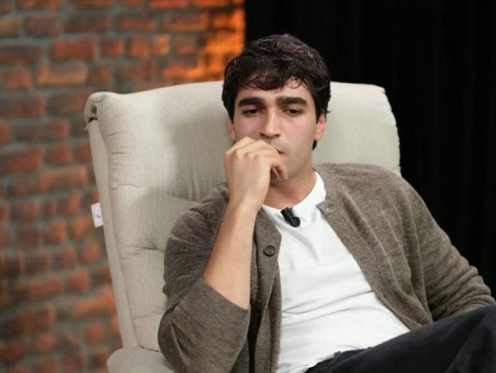 Mert Ramazan Demir poartă un tricou alb și stă pe un scaun pe un fundal deschis la culoare